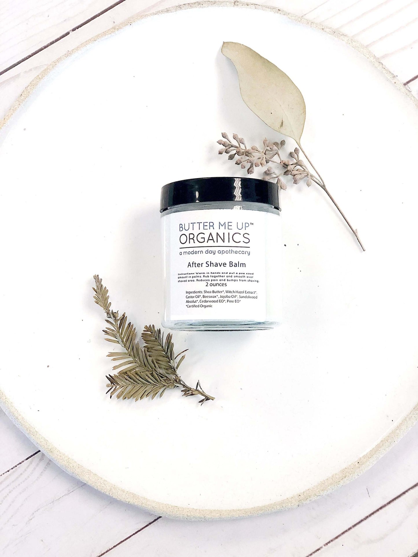 Organic After Shave Balm in clear jar with black lid, surrounded by herbs on a white plate.