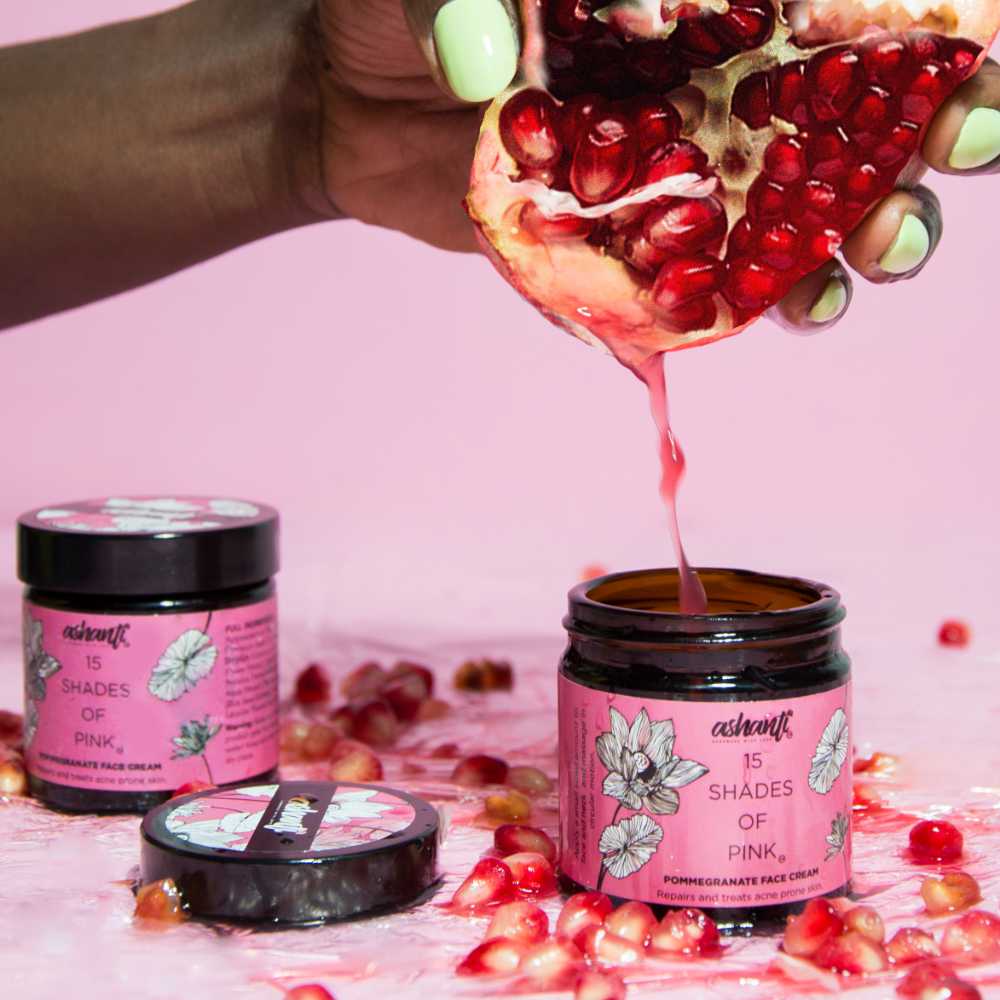 Opened pomegranate spilling juice over Ashvant's 15 Shades of Pink Pomegranate Face Cream jars on a pink background.